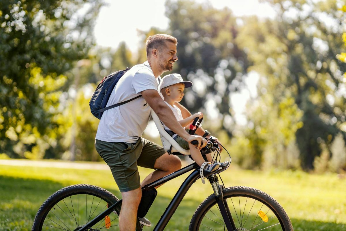 A man rides a bicycle in a park with a young child sitting in a front child seat on a sunny day.